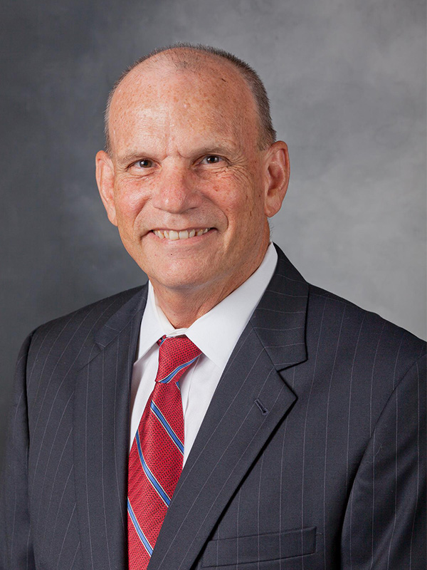 A headshot of a man wearing a navy blue suit, white shirt, and red tie.
