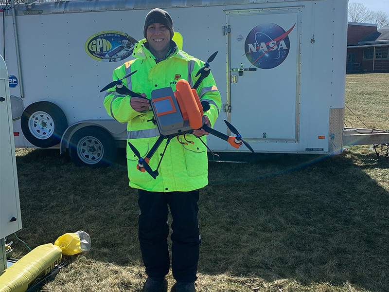 A person holding a drone, wearing a high visibility jacket and standing in front of a trailer with a NASA logo on it.