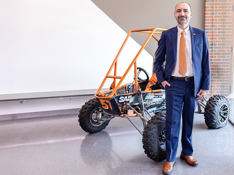 A man in a navy blue suit and orange tie stands in front of a formula race car.