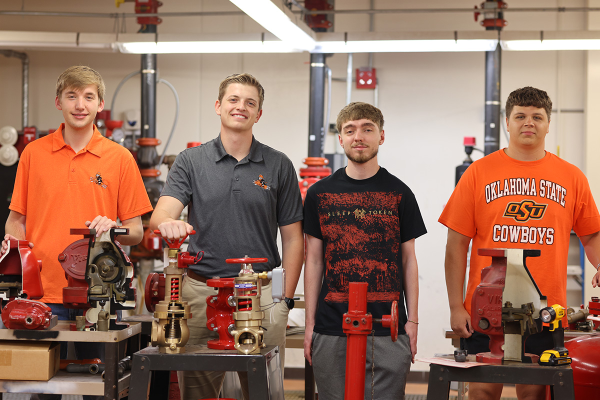 Four students stand in front of fire sprinkler equipment, dressed in OSU apparel, in a lab.