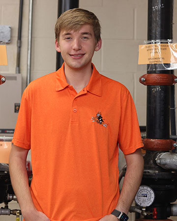 A student stands in a fire sprinkler lab dressed in OSU orange polo.