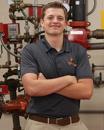 A young man stands in front of a sprinkler system dressed in business casual attire.