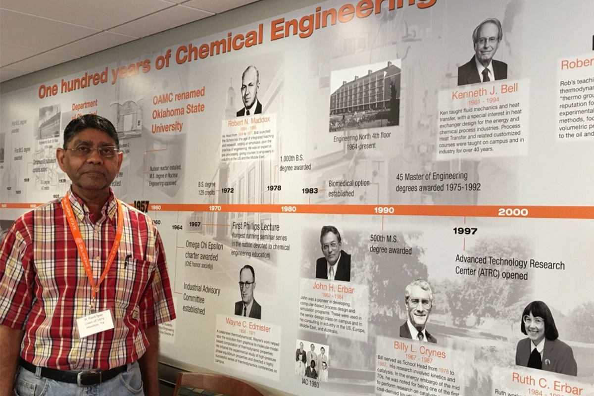 A man dressed in a red plaid shirt stands infront of a timeline depicting the 100 year history of OSU's School of Chemical Engineering.