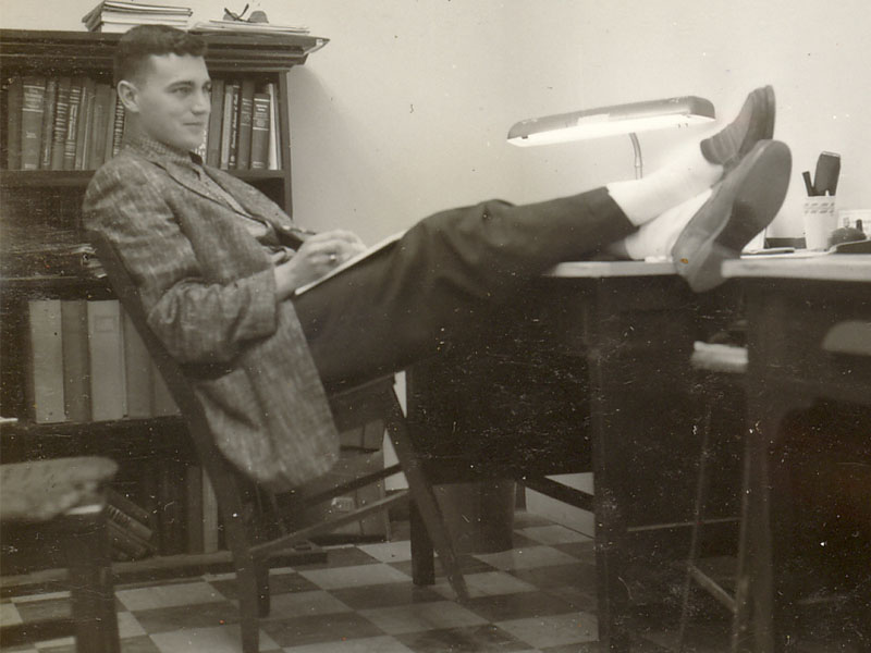 A young man in professional clothing leans back in a chair with his feet up on a desk.