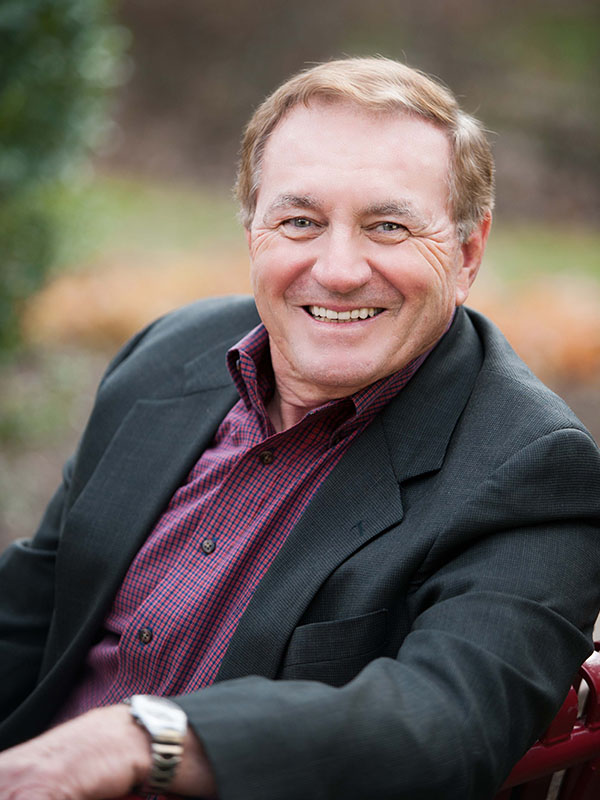 A professional headshot of a man in a black suit and purple dress shirt.
