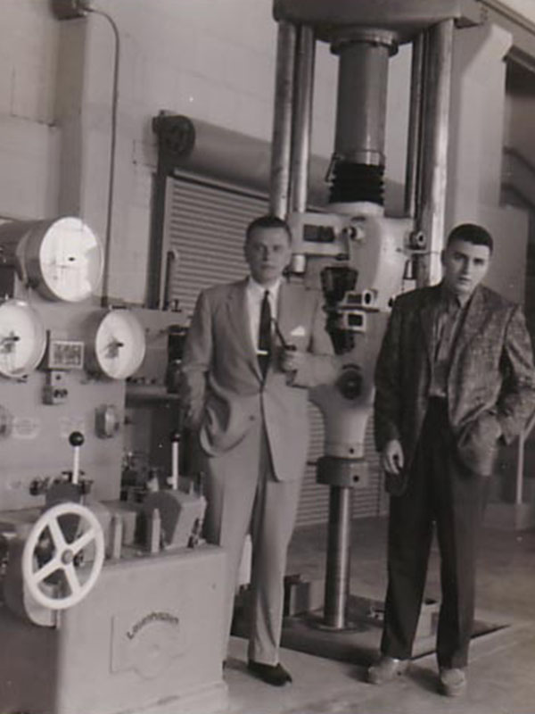 Two men in suits pose near a large piece of equipment in a laboratory.