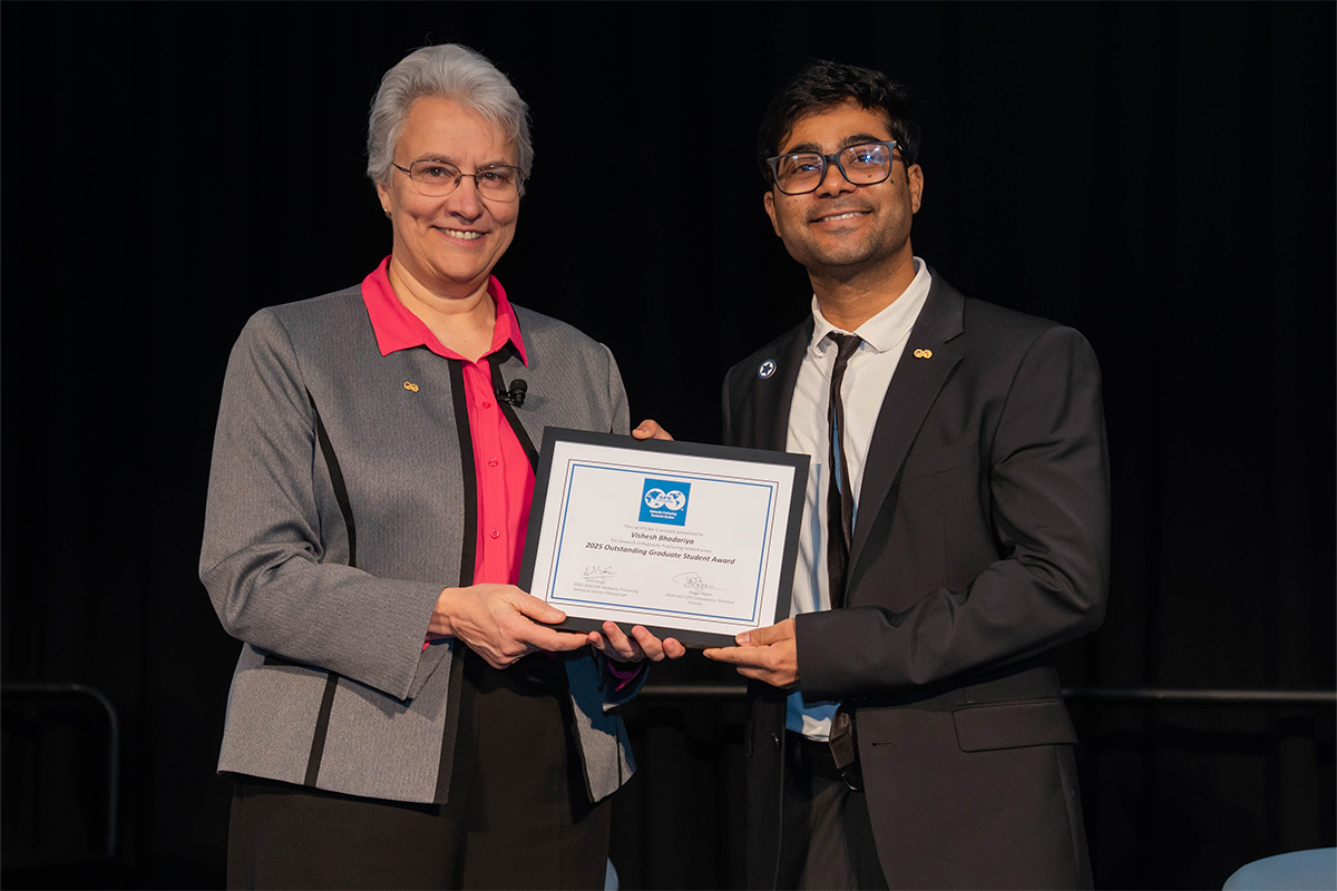 A man and woman stand on stage in business professional attire. The man is holding a certificate.
