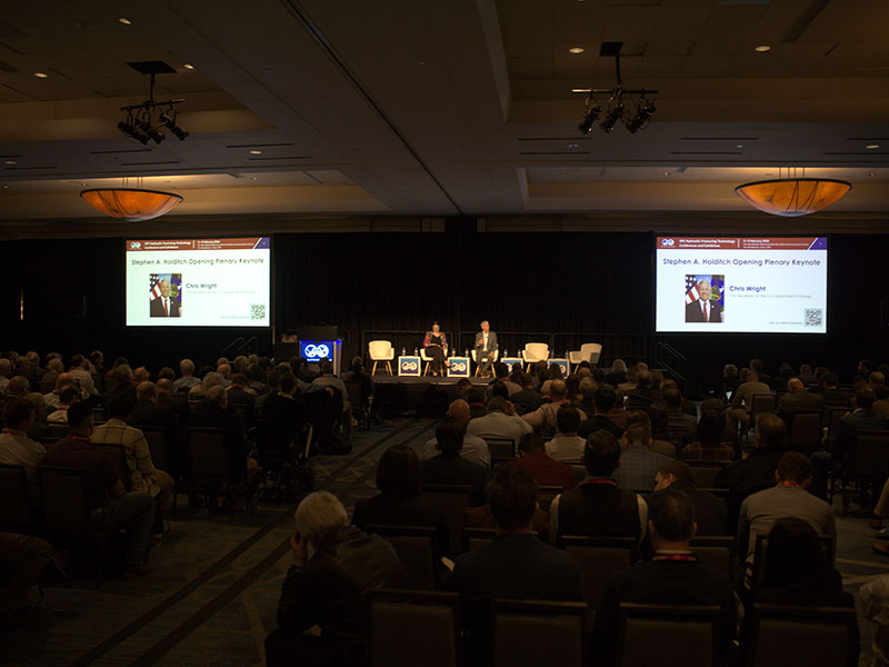 A picture of a large room filled with individuals listeneing to two people on stage discuss a topic.