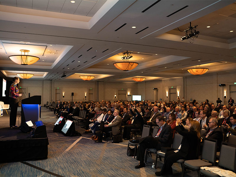 A side angle view of a conference room filled with people in chairs and a stage with two projectors at the front of the room.