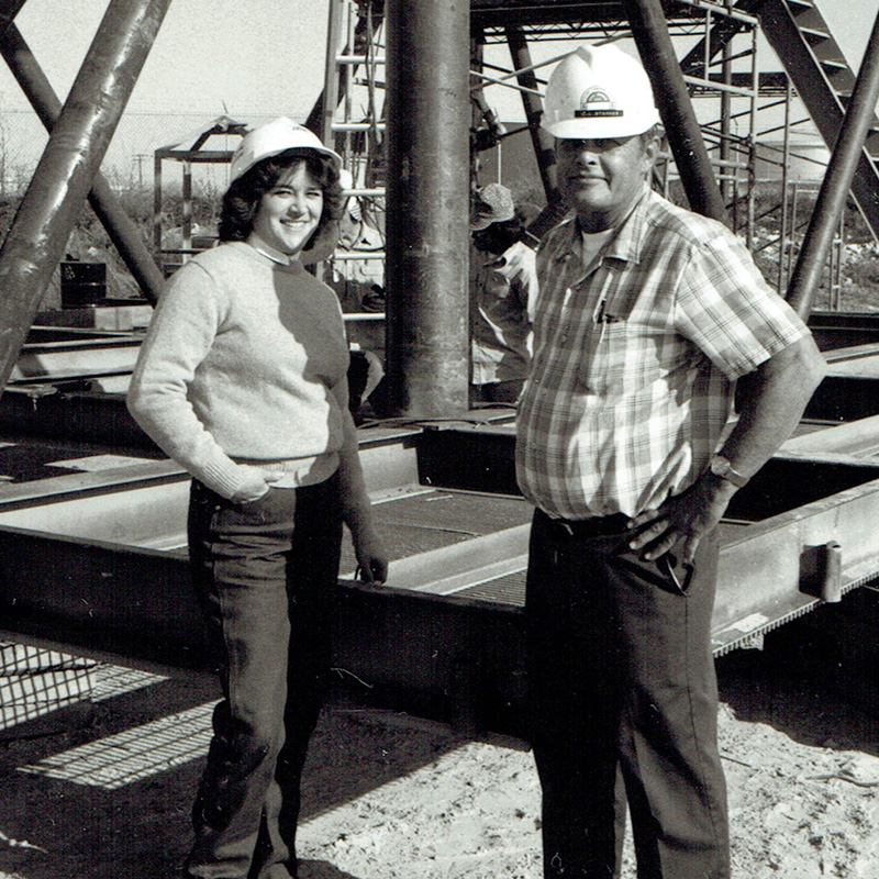 A black and white photo that features a man and a woman in hard hats standing infront of oil equipment. 