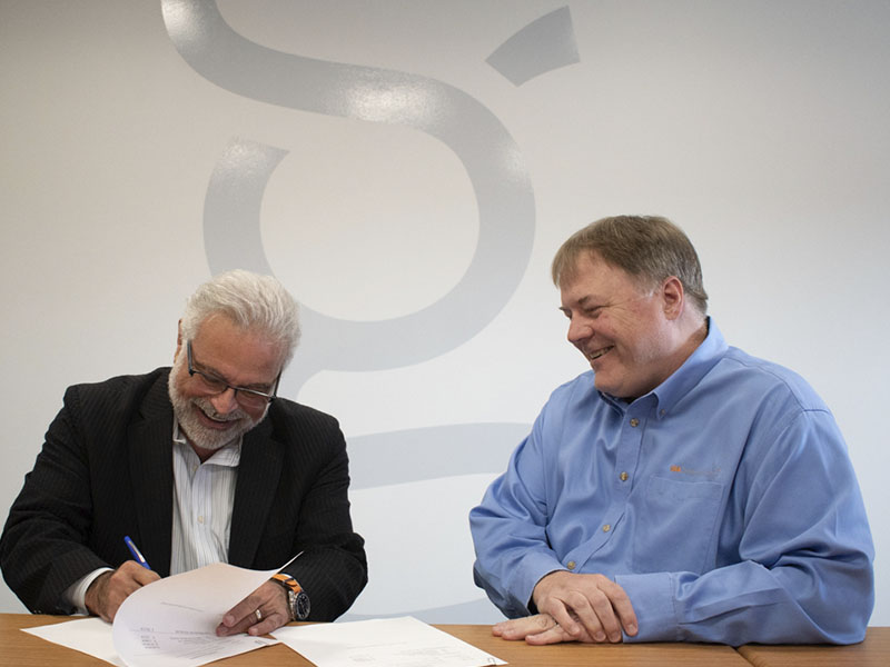 Two men sit at a large table signing legal paperwork