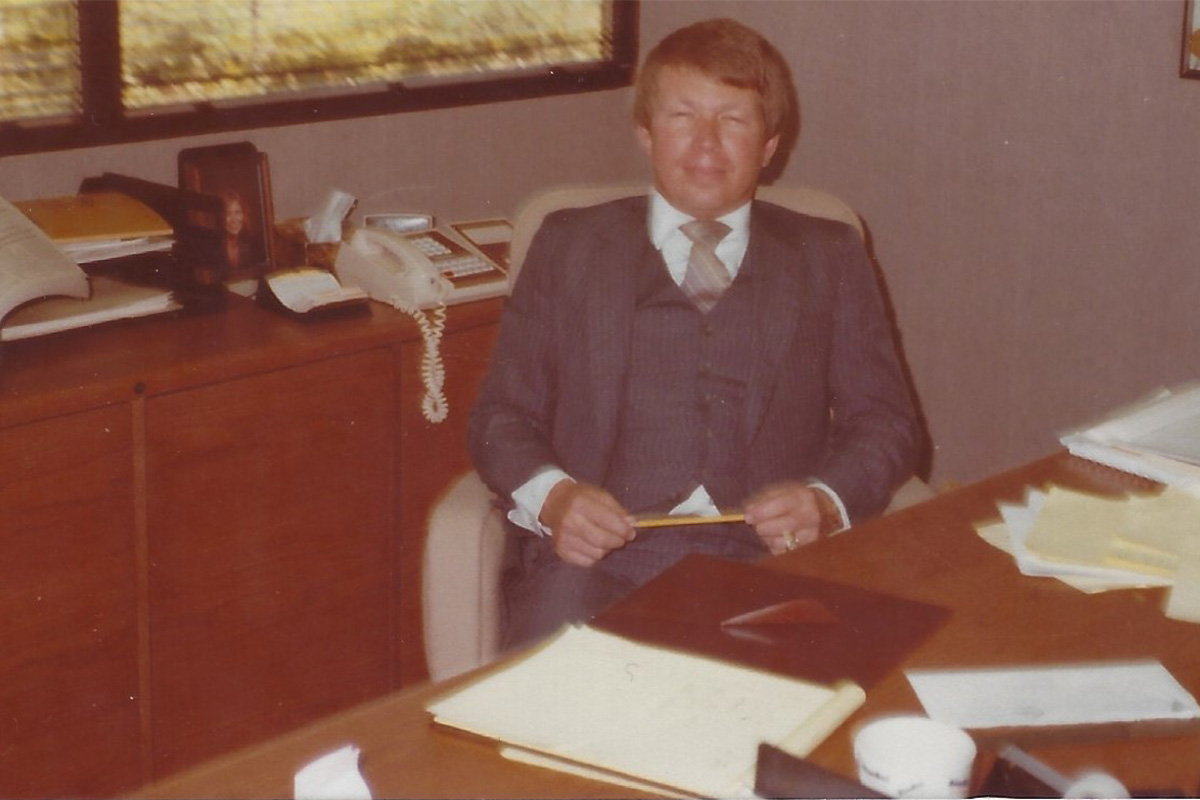 A photo of a man sitting at his desk in a grey suit. 