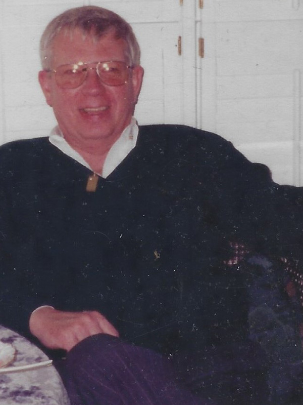 A smiling man sitting on a white couch in a navy blue sweater.