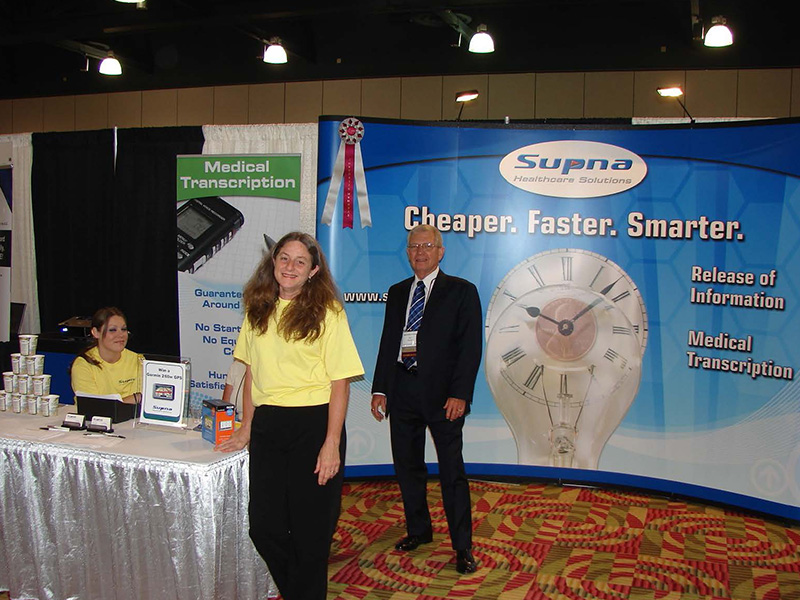 Two ladies in yellow shirts stand near a booth at a convention with a man in a black suit standing in front of the company's banner. The banner states, 