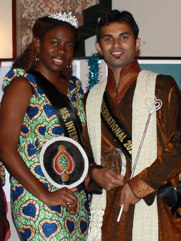 A man and woman dressed in traditional cultural wear pose together holding awards and wearing sashes that state their title.