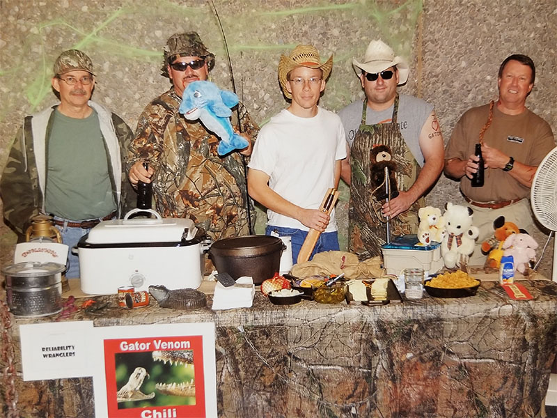 A group standing behind a table decorated with camo and fishing gear prepared to serve chili. 