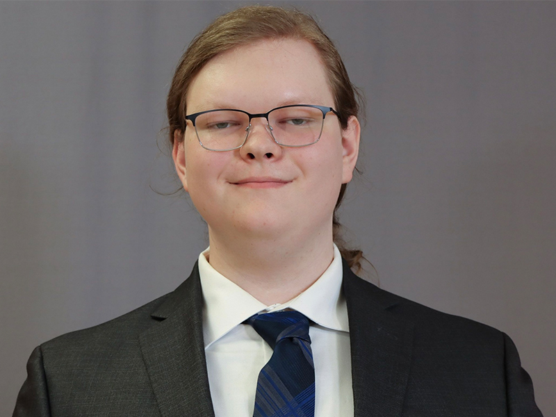 headshot image of Collin Stringer in a black suit, navy/gray tie standing in front of a gray backdrop